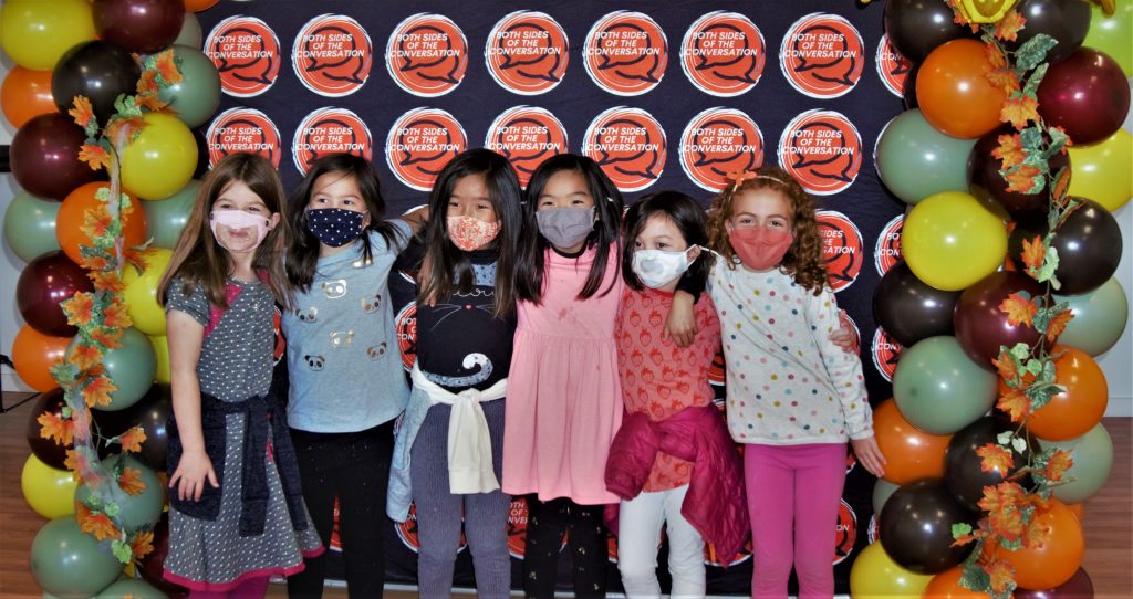 A group of diverse girls wearing face masks stand in front of a colorful banner and smile.