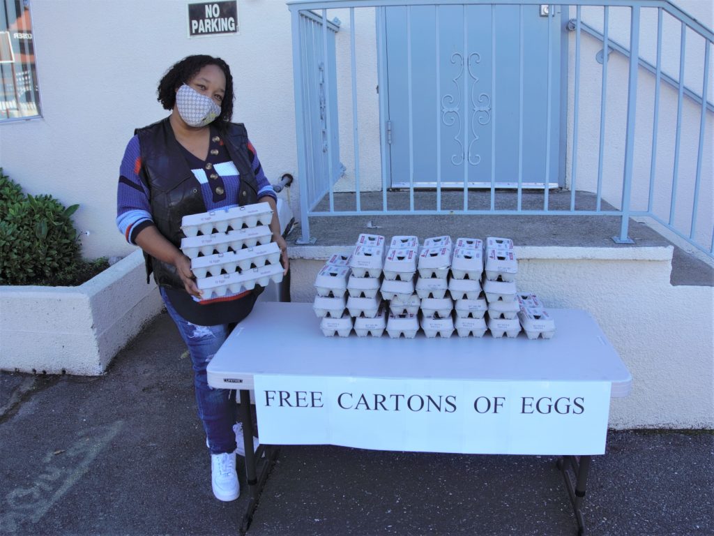An African American woman stands by a table filled with egg cartons and a "free eggs" sign.