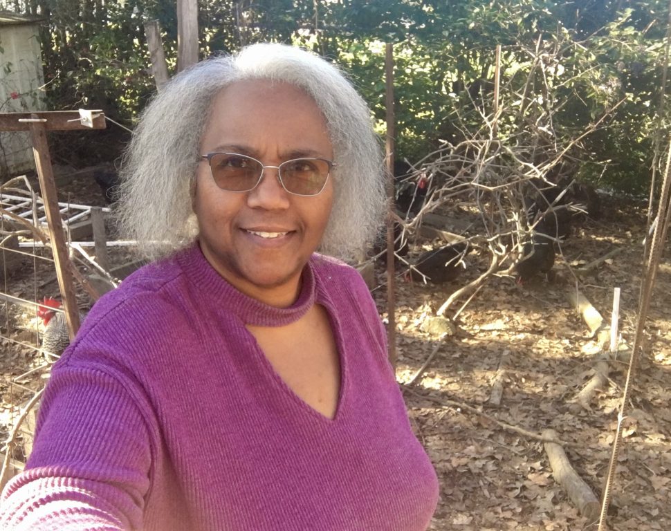An older Black woman with grey hair wearing a purple shirt stands in front of a garden bed holding a rake.