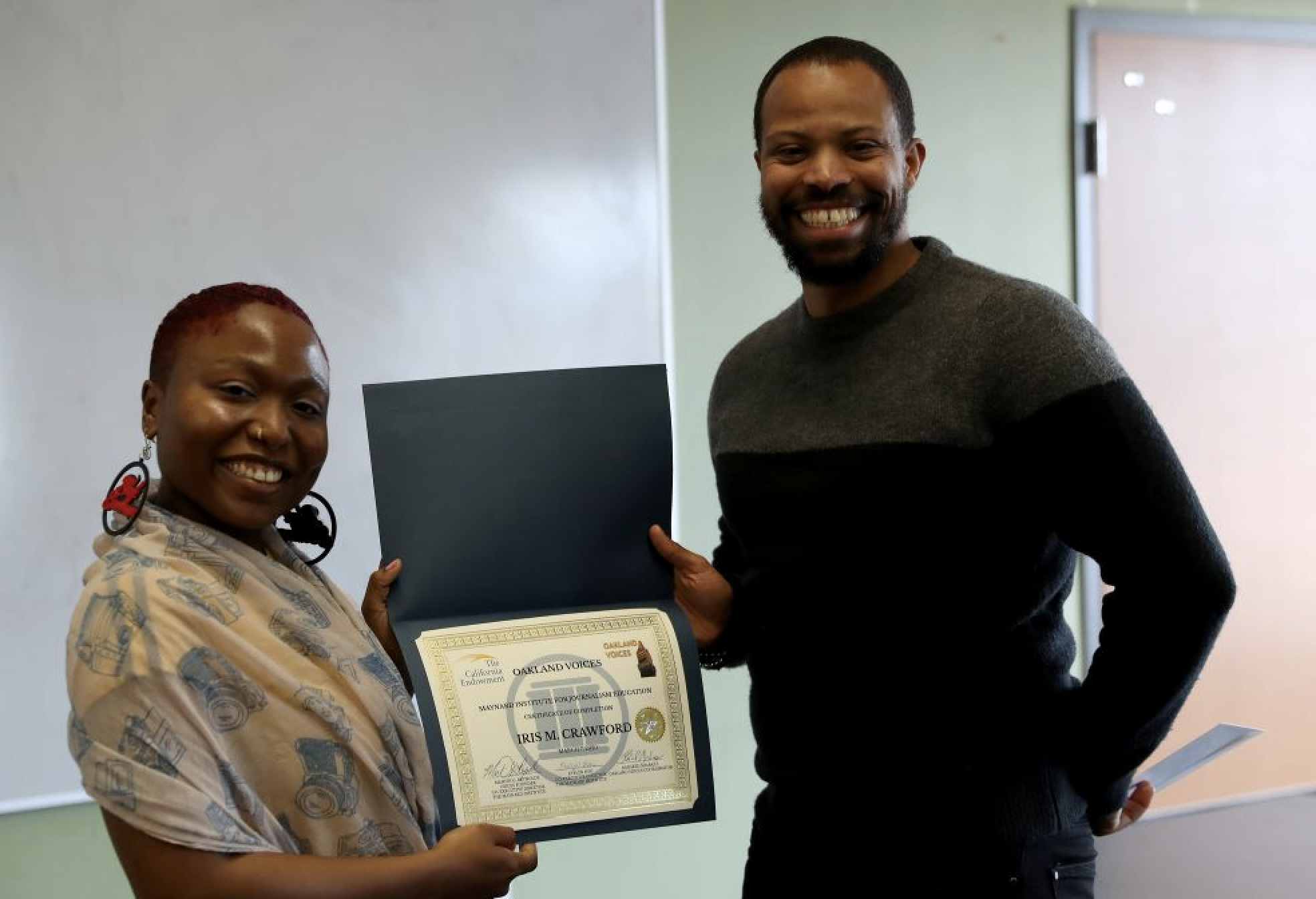 An African American woman with short hair holds a diploma with a taller Black man and they are both smiling.