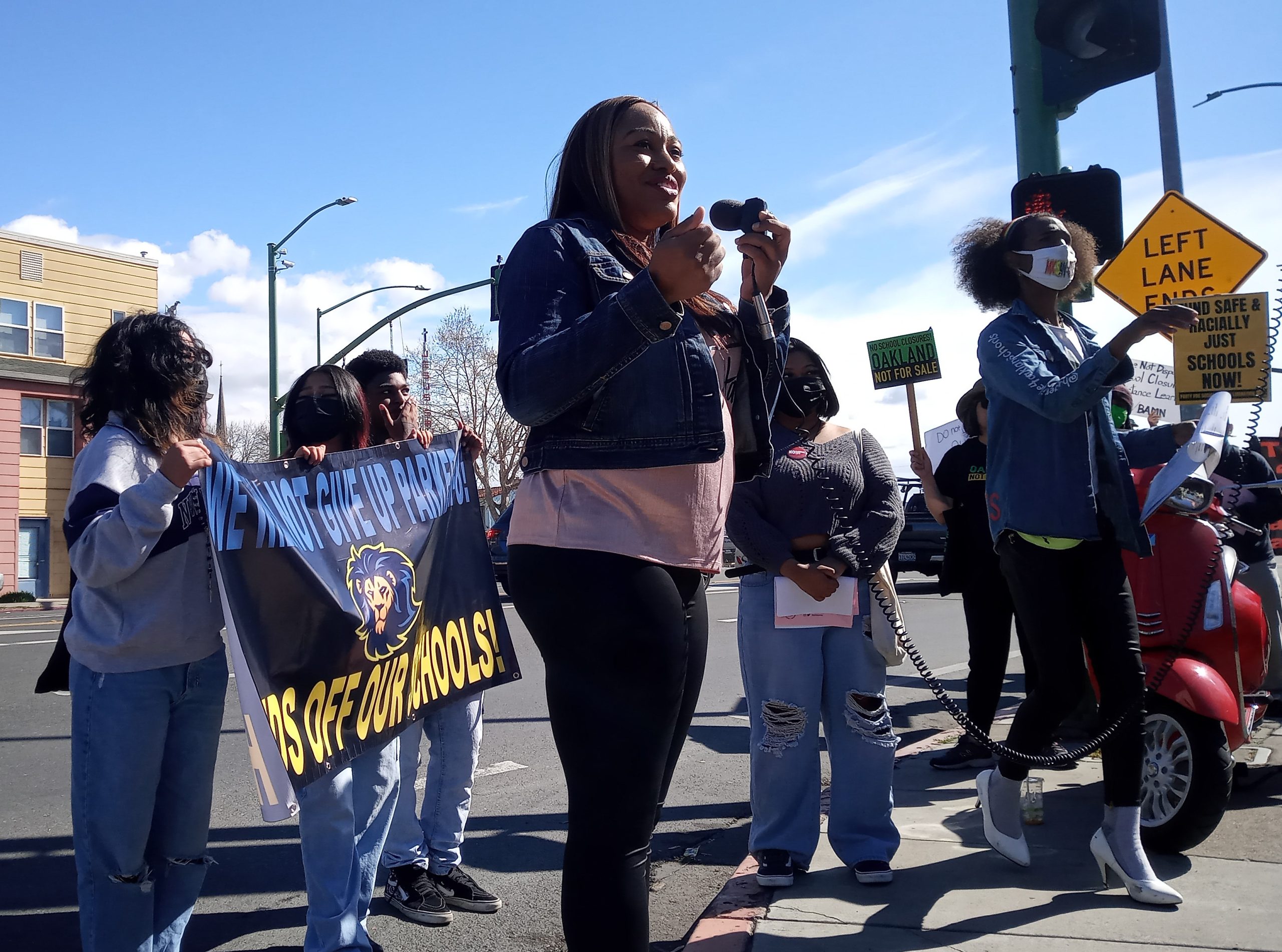 A black woman with long hair speaks into a mic outdoors in front of a school. with students standing behind her