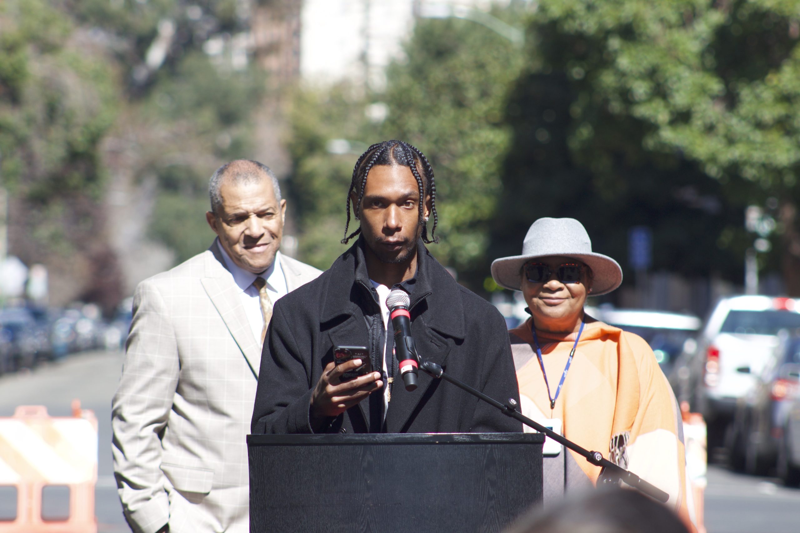 A Black man in an off white suit, a young Black man wearing a black jacket, and Black woman with sunglasses, smiling, in grey hat stand outside in front of podium