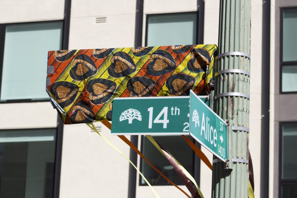 two green street signs state 14th and Alice and another sign covered by traditional Mali cloth