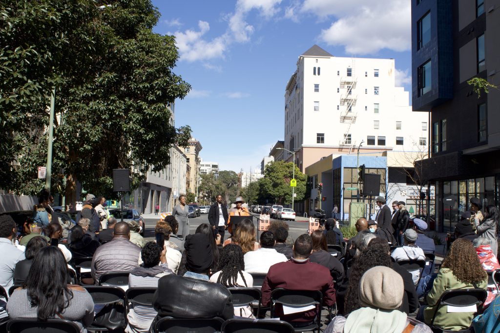 A crowd sits facing a street while an African American woman speaks at the front 