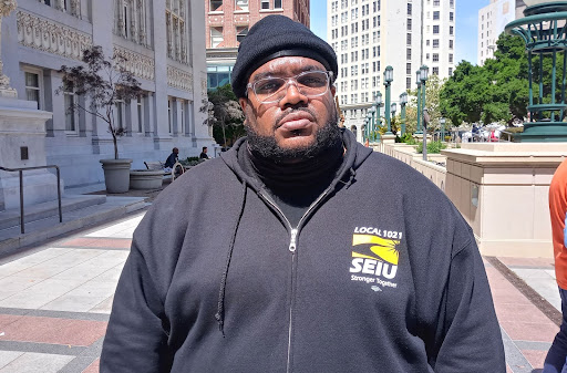 A Black man wearing a black beanie and glasses also wearing a hoodie that says seiu local 1021 poses for camera
