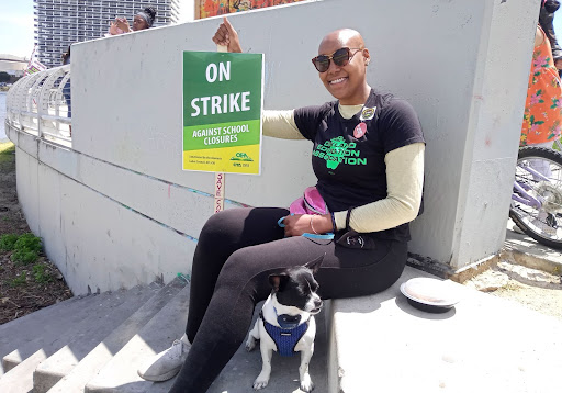 A Black woman with bald hair wears a black t-shirt that says OEA and holds green sign that says ON STRIKE