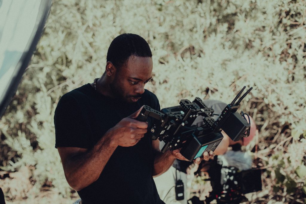 An African American man with short hair directs film behind a video camera