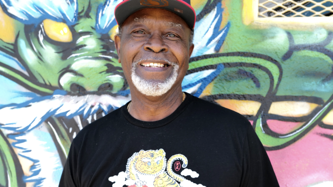 An elder African American man wears Warriors cap and black t-shirt with a lion/dragon image on it, standing in front of bright mural.