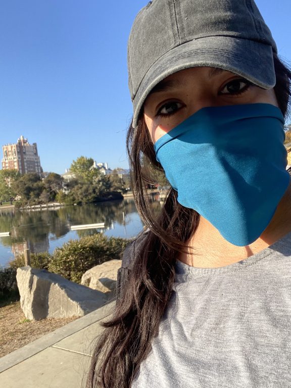 Woman wearing grey shirt and hat, with blue mask, stands next to Lake Merritt.