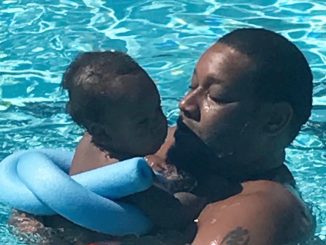 A Black man holds his infant son in the water with a blue pool noodle tied safely around the baby to keep him afloat
