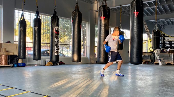 A boxing gym in East Oakland with concrete floors and bags handing from ceiling while one man is boxing in the right corner
