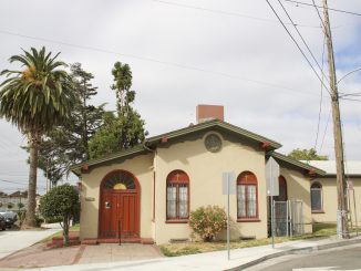 A beige and brick colored small quaint church next to palm tree and electric lines, with gold painted fan details on the doorway and a gold cross on the front of church
