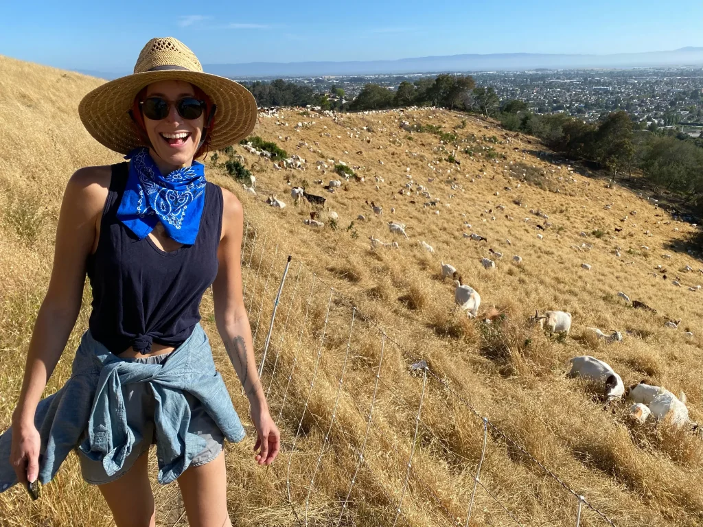A woman wearing a straw hat and blue tank top smiles on a hike in the hills