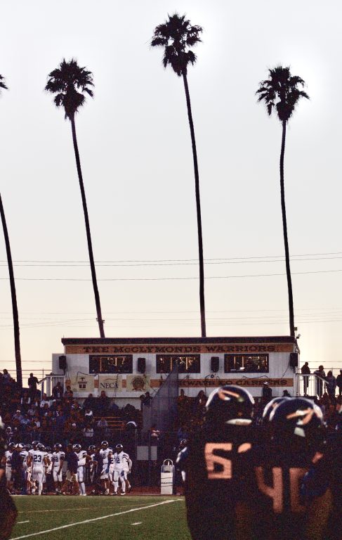 bleachers and clubhouse that says "McClymonds' with sun setting in back and three tall palm trees in the back