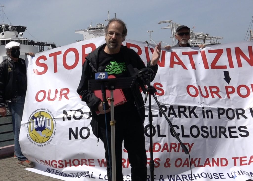 An older white man wearing all black stands in front of large banner that says "stop the privatization of our public schools" while he speaks into a mic