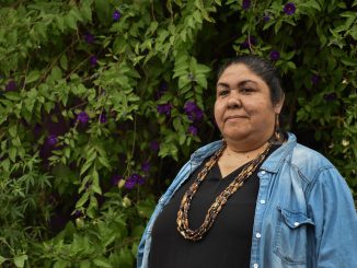 An Indigenous woman wearing denim and beaded necklace stands in front of green trees