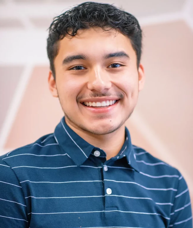 A young Latino man wearing blue striped polo shirt smiles for head shot