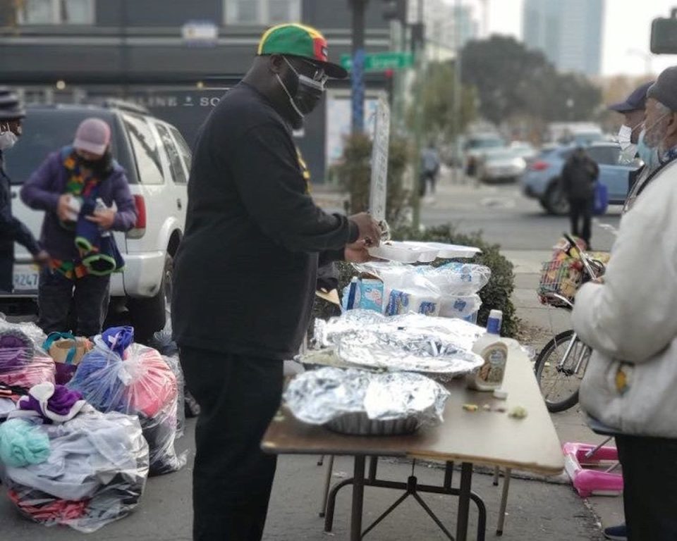 An African American man with green, yellow, and red camp stands beside portable outdoor table handing out food