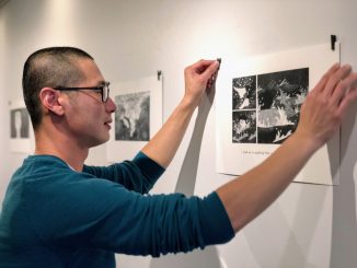 An Asian American man with a shaved head and glasses waring teal sweater puts up a black and white art print on the wall