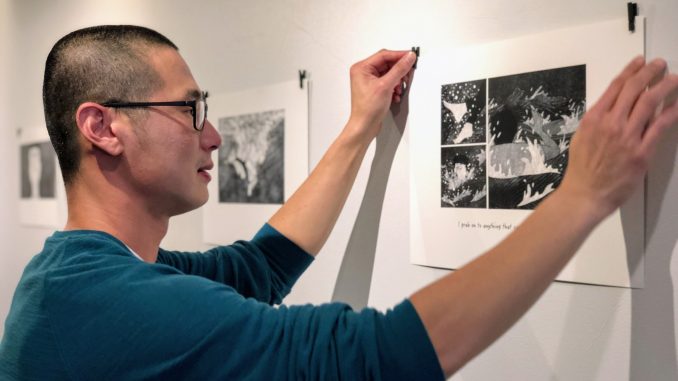 An Asian American man with a shaved head and glasses waring teal sweater puts up a black and white art print on the wall