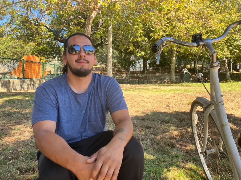 A man wearing shades and blue t-shirt and nose piercing sits at park with his bike