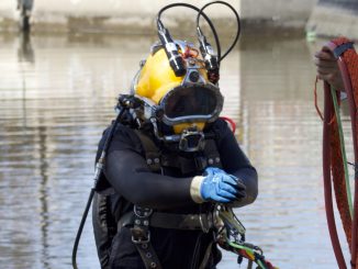 A man in a wetsuit and a yellow hard helmet comes out of murky waters