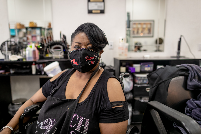 An African American woman with short hair and wearing face mask sits inside hair salon