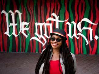 An African American woman wearing a black hat and shades and red shirt and black jacket stands in front of a colorful mural that says "we got us"