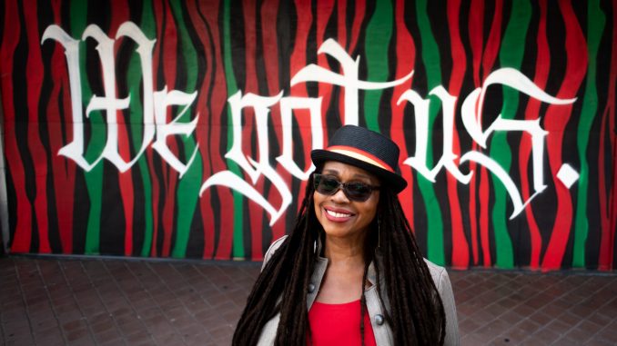 An African American woman wearing a black hat and shades and red shirt and black jacket stands in front of a colorful mural that says "we got us"