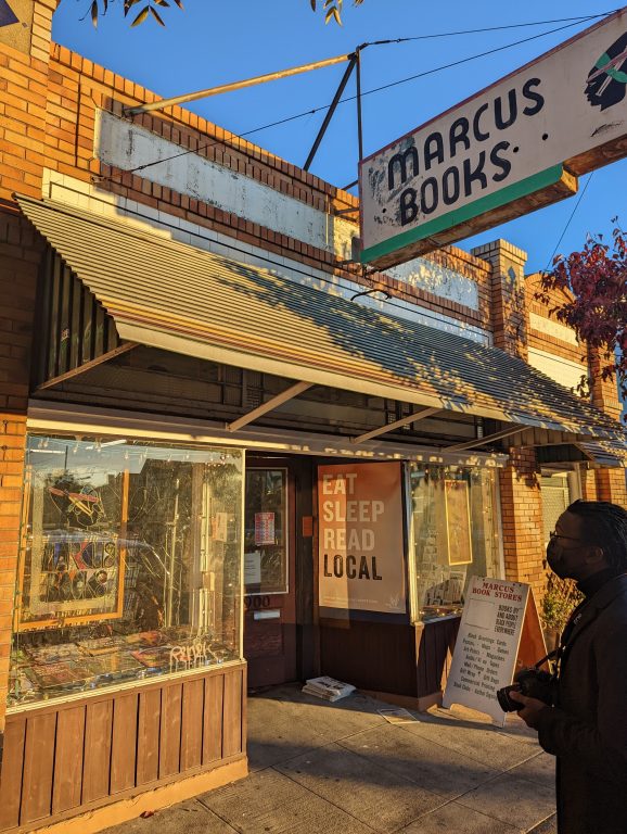 Outside a small bookstore with sun gleaming on the sign that says Marcus Books