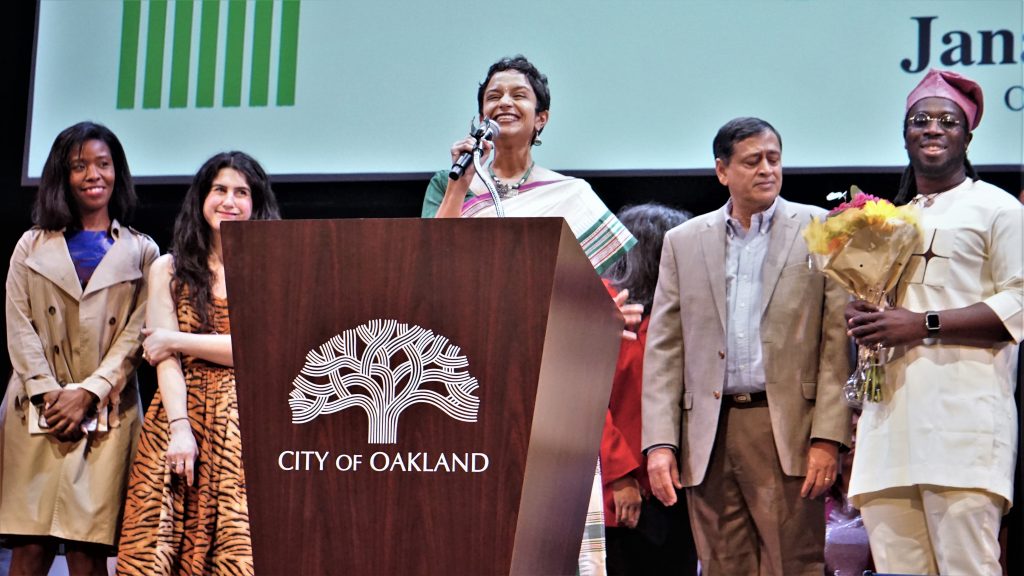 A South Asian woman with a pixie cut wearing a sari stands behind podium speaking with a smile, while diverse family and friends stand near her