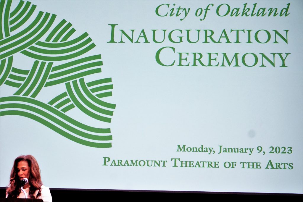 A Black woman with brown hair is in lower bottom left of image, with a large projection behind her that says "City of Oakland Inauguration Ceremony"