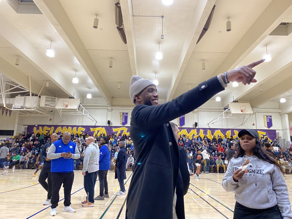 A smiling African American man wearing a grey beanie points to crowd inside a basketball gymnasium