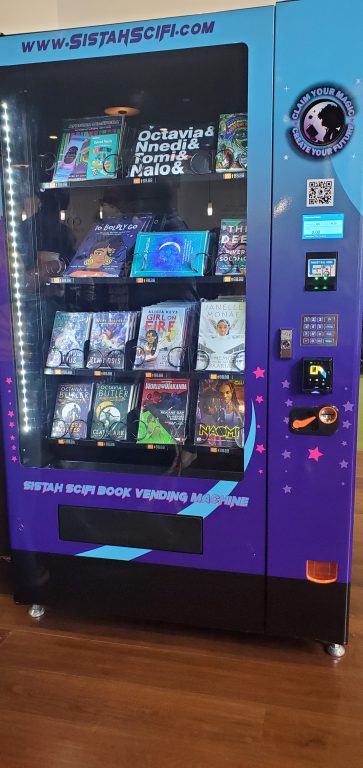 a purple vendor machine contains books and a t-shirt of Black and Indigenous Afroscifi works