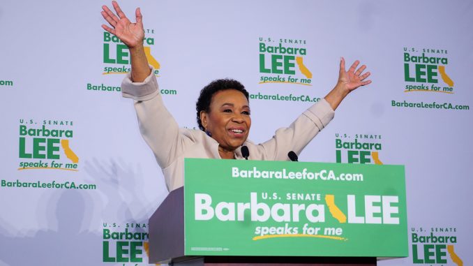 An African American woman with short hair raises her hand in celebration standing behind a podium. The sign in front says "Barbara Lee" in a green poster