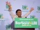 An African American woman with short hair raises her hand in celebration standing behind a podium. The sign in front says "Barbara Lee" in a green poster