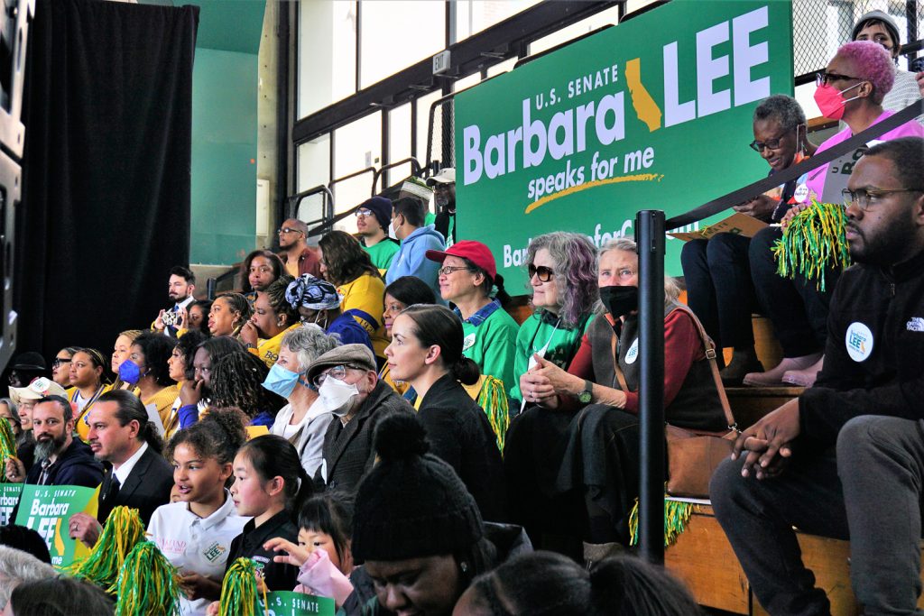 A throng of diverse people wearing yellow and green t-shirts supporting Barbara Lee in the stands