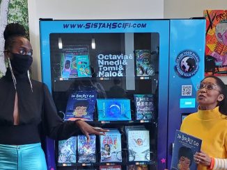 A Black woman wearing teal pants and black face mask stands in front of colorful vending machine, with a shorter woman wearing yellow t-shirt and glasses holding a sci-fi book she wrote