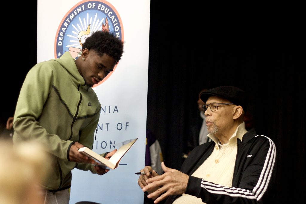 An African American boy holds a book up for an elder African American man to sign the book.