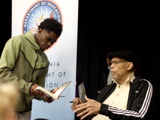 An African American boy holds a book up for an elder African American man to sign the book.