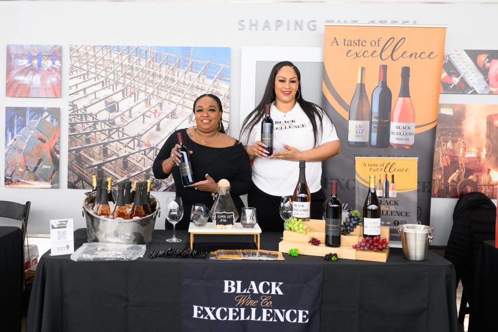 Two African American women hold up bottles of wine at a table showcasing local wine