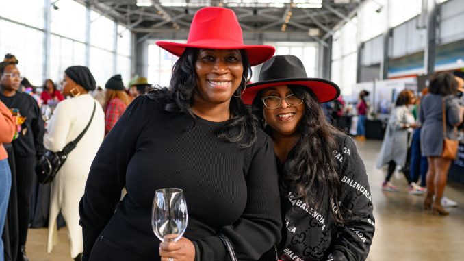 Two African American women wearing nice hats hold a wine glass and smile at an indoor spacious Black wine festival in Oakland