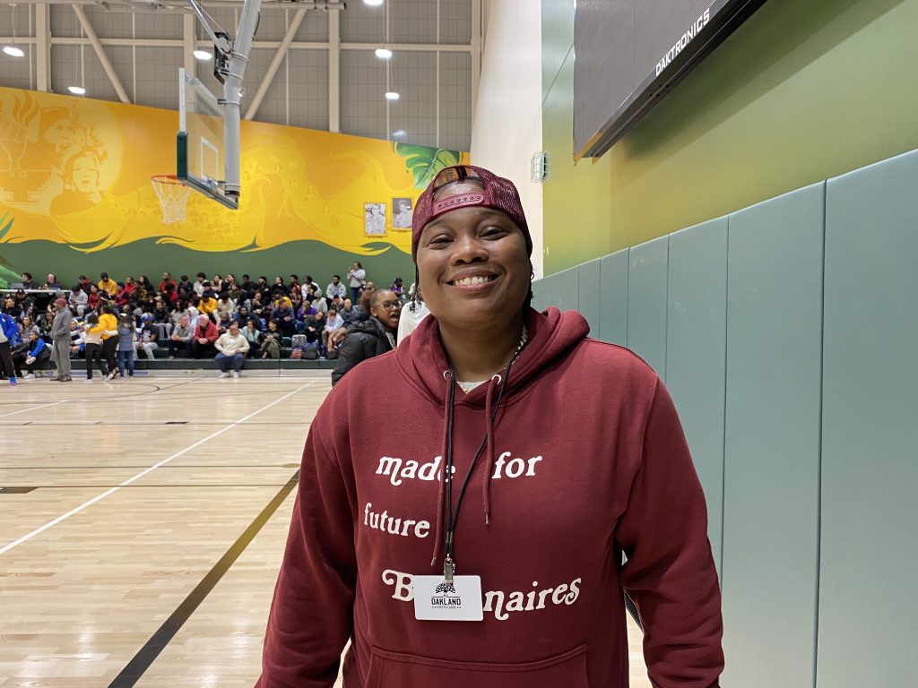 An African American coach/leader wearing maroon stands inside colorful gym and wearing a baseball cap backwards