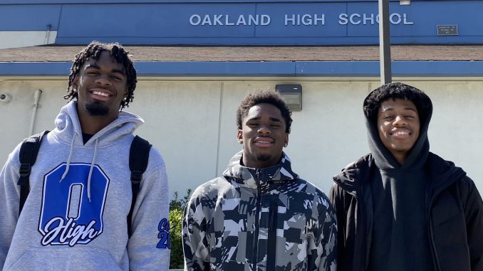 Three African American boys wearing sports gear and hoodies stand in front of their school, Oakland High, and smile at camera