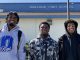 Three African American boys wearing sports gear and hoodies stand in front of their school, Oakland High, and smile at camera