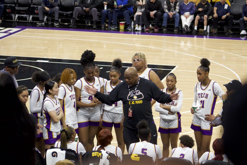 A group of mostly African American girls, with one Asian girl, huddle around a Black male coach during a time out in a championship game