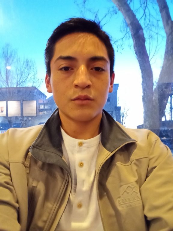 A young Latino man wearing a khaki jacket takes selfie