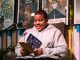 An African American boy sits on stage reading from a book he wrote with a smile on his face