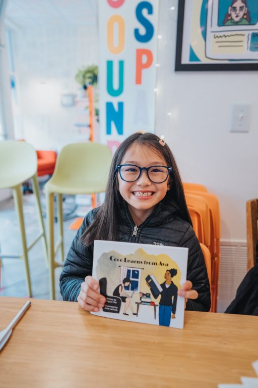 A young girl wearing glasses holds up a book she wrote