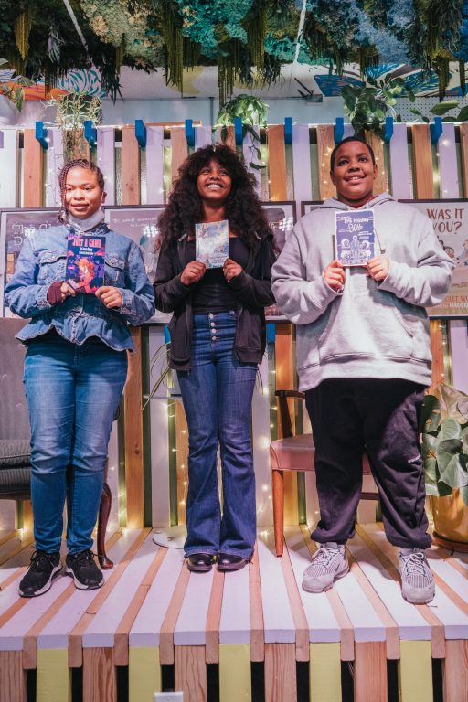 three African American youth stand on a stage holding books they wrote